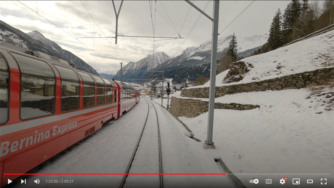 photograph from YouTube of a red Bernina Express passenger train traveling through snow towards mountains in Switzerland.