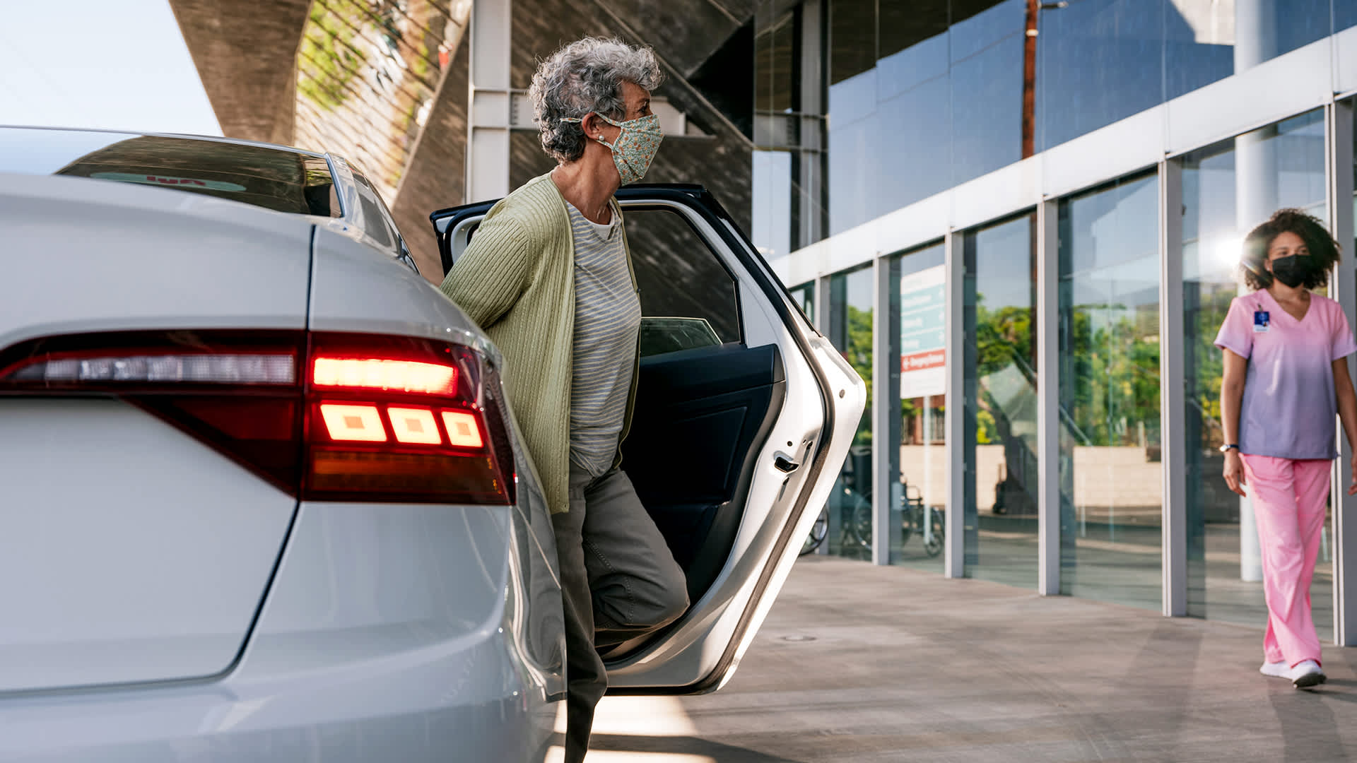 A photograph of a senior woman, wearing a face mask, exiting a car in front of glass doors at a medical facility.