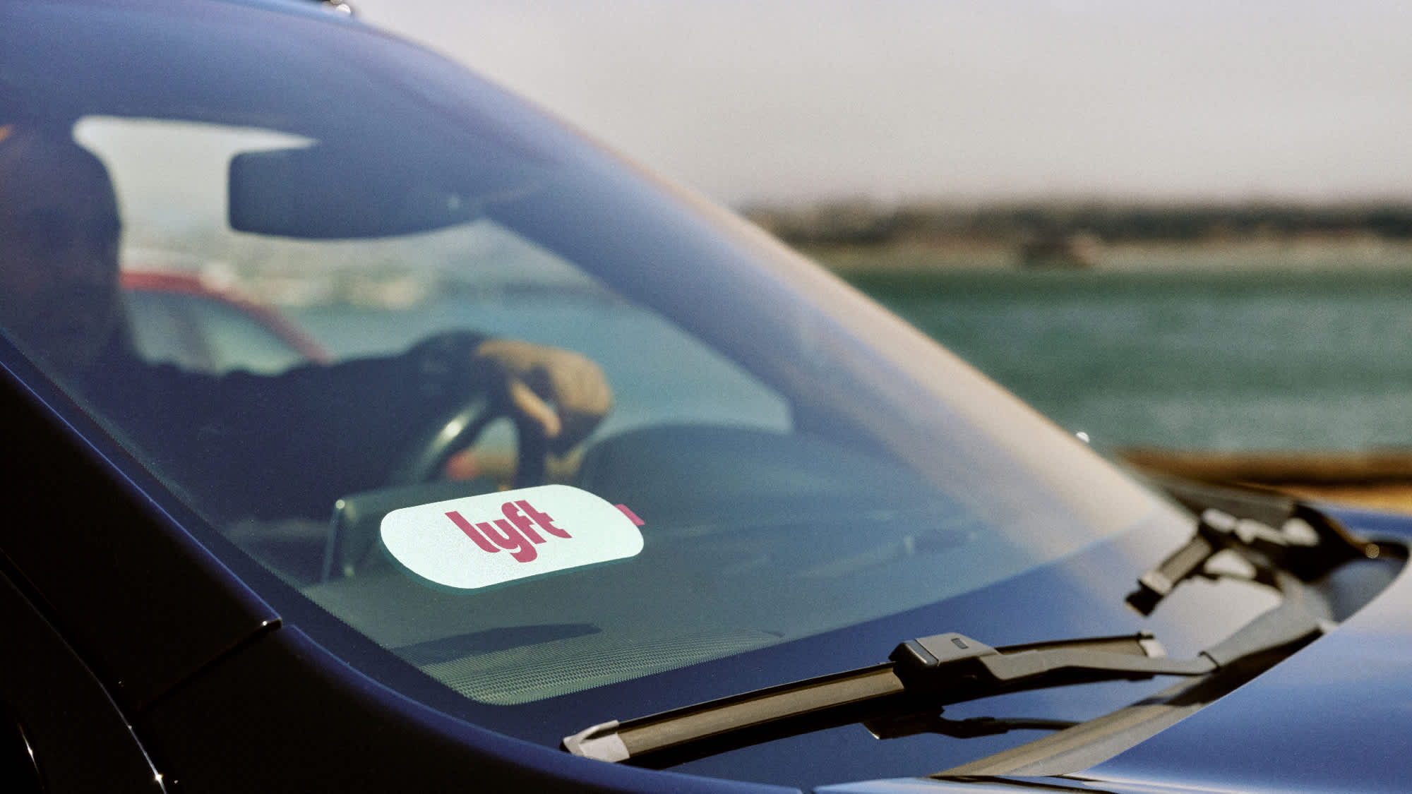 Car windshield with Lyft logo sticker, driver visible, with water in the background. 
