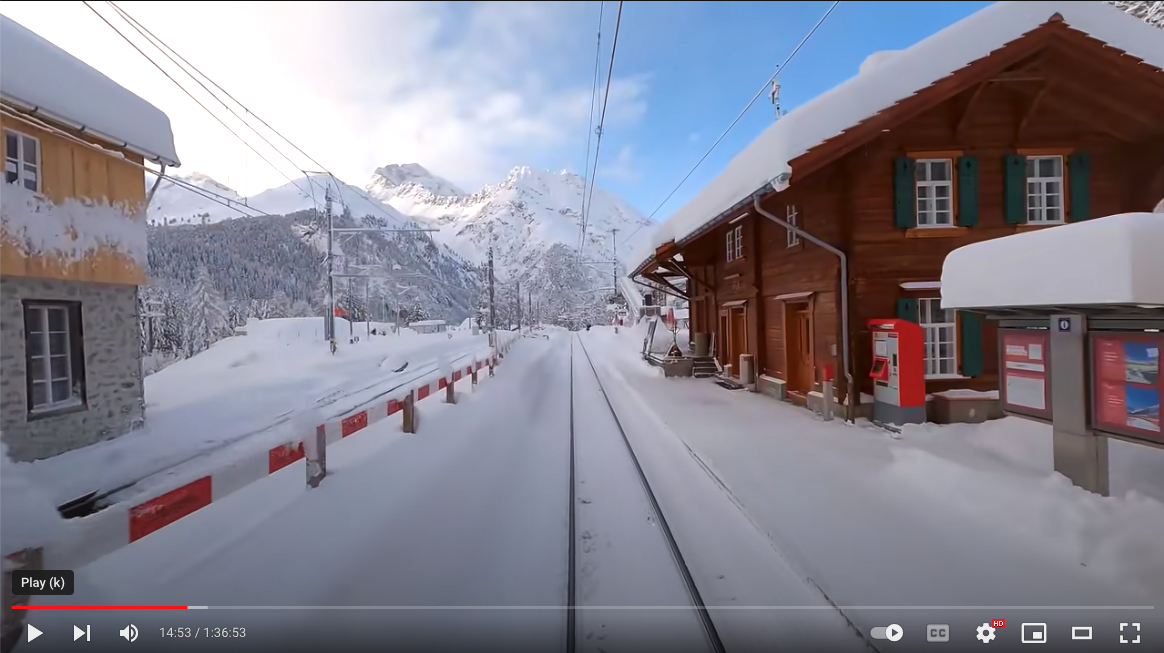 Image from Youtube of a snowy train station, tracks and mountains in Switzerland.