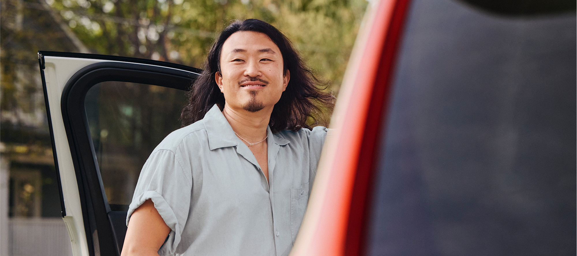 Person with long dark hair in light blue shirt smiling beside open car door outdoors.