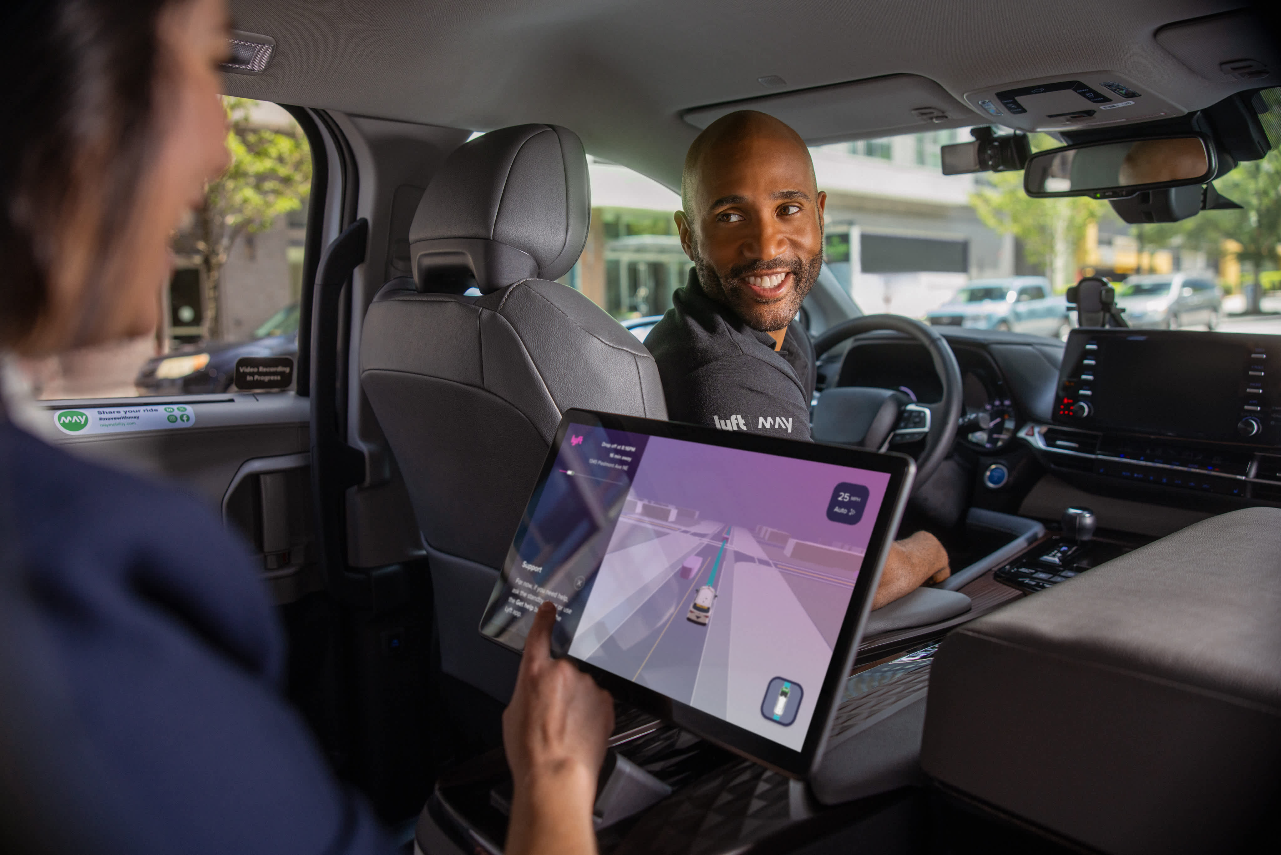 Smiling driver turning to passenger holding tablet with navigation app in vehicle interior.