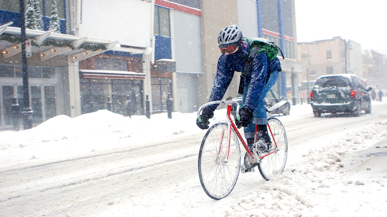 Photograph of a cyclist wearing a helmet, ski goggles and gloves riding a red road bike on a snow covered street.