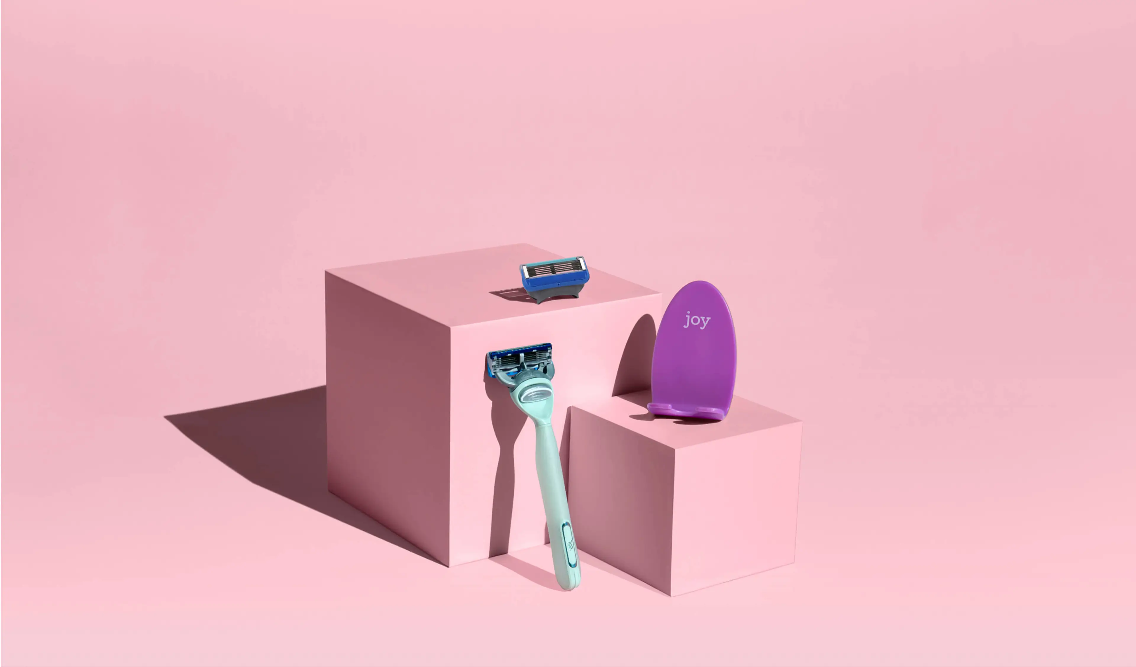 Rasoir Joy, recharge de lame et accessoire de rasage violet présentés sur des blocs roses.