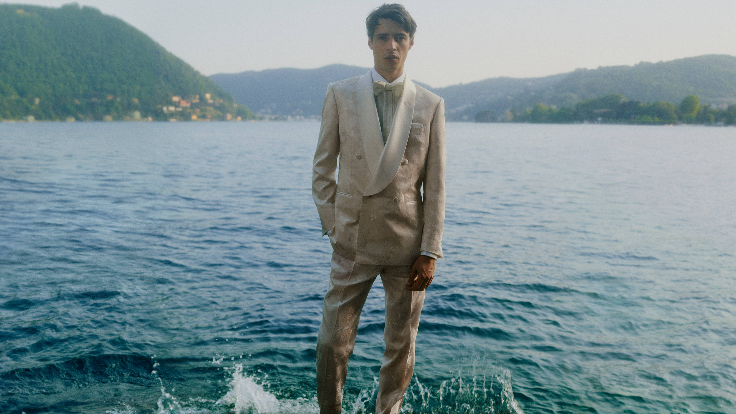 Model wearing a light beige double-breasted Brioni suit in a lakeside landscape.
