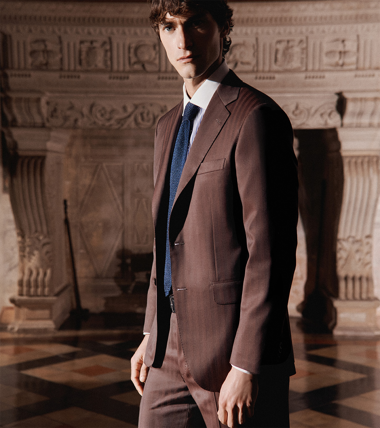Model wearing a brown pinstriped Brioni suit in an elegant interior.