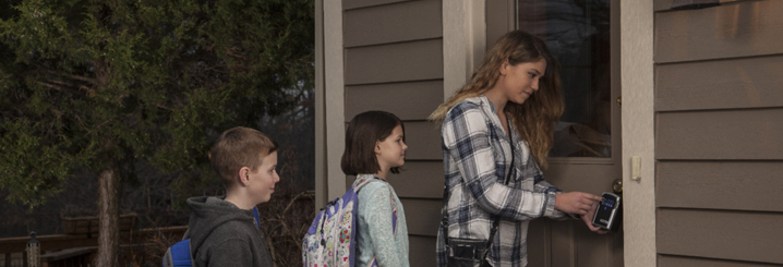 A baby sitter uses a lock box to retrieve a key to the front door of a home. Two younger children are with her with backpacks on waiting patiently. 