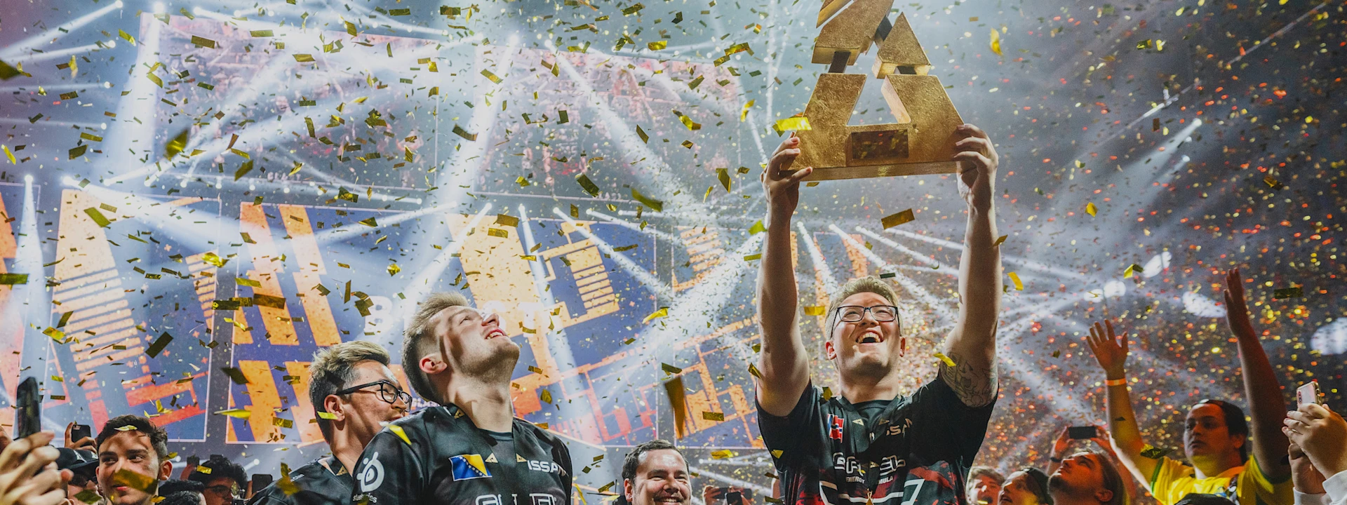  Two victorious esports players celebrate on a crowded stage under a shower of golden confetti, one joyfully hoisting a large, golden trophy.