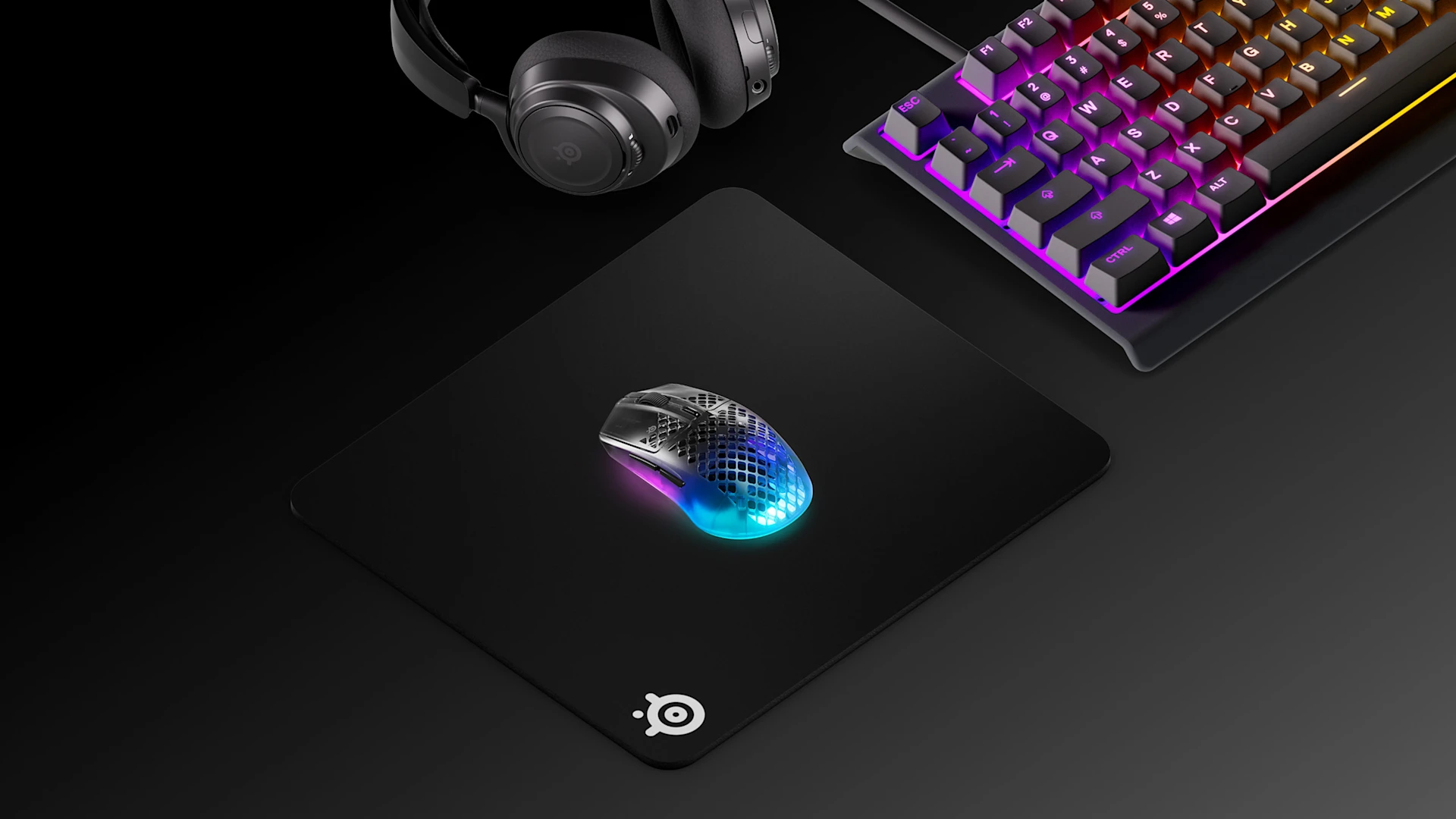Widok izometryczny klasycznego czarnego zestawu gamingowego SteelSeries, w skład którego wchodzą podkładka pod mysz QcK Heavy L, klawiatura Apex RGB, zestaw słuchawkowy Arctis Nova oraz mysz Aerox 3 Wireless.