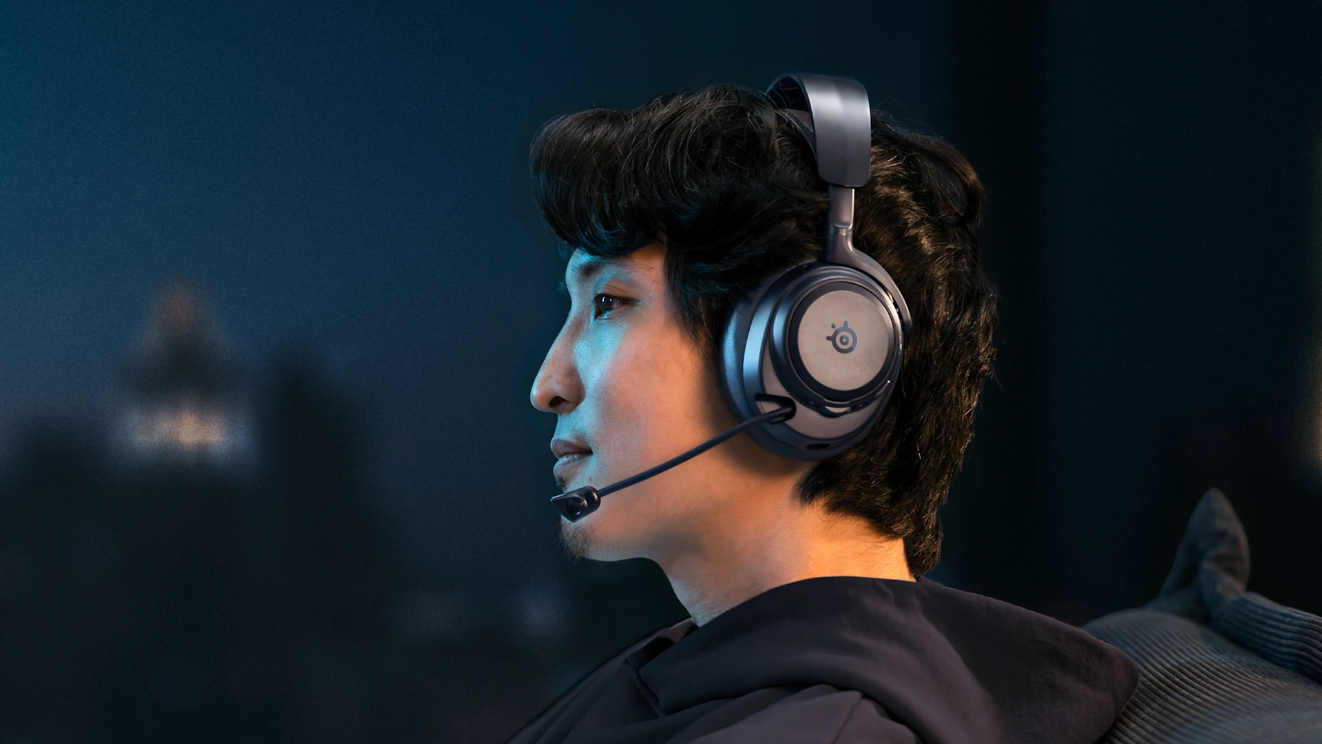 Osoba nosící náhlavní sluchátka SteelSeries, která večer relaxuje doma, s rozmazaným výhledem na město a jemným modrým osvětlením v pozadí.