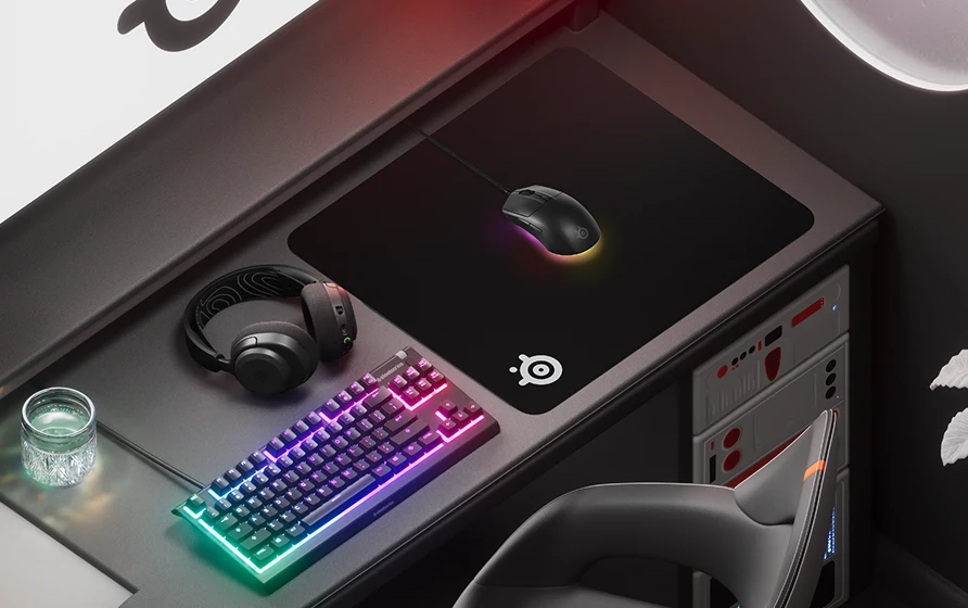 Una configuración para juegos en tonos negros con una alfombrilla SteelSeries, un ratón con un cálido brillo RGB, auriculares a juego y un teclado RGB compacto.