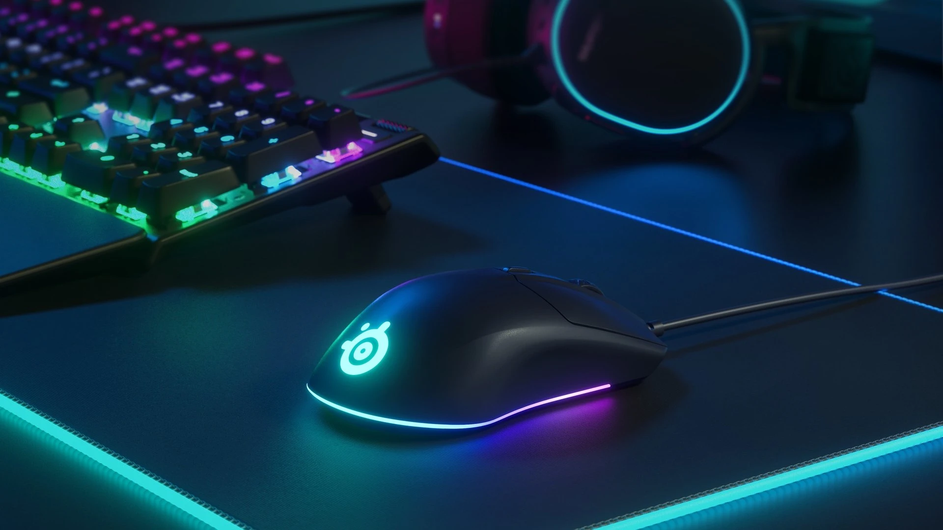 Rival 3 wired mice with other SteelSeries gears: RGB keyboard, RGB surface and RGB headset in the background.