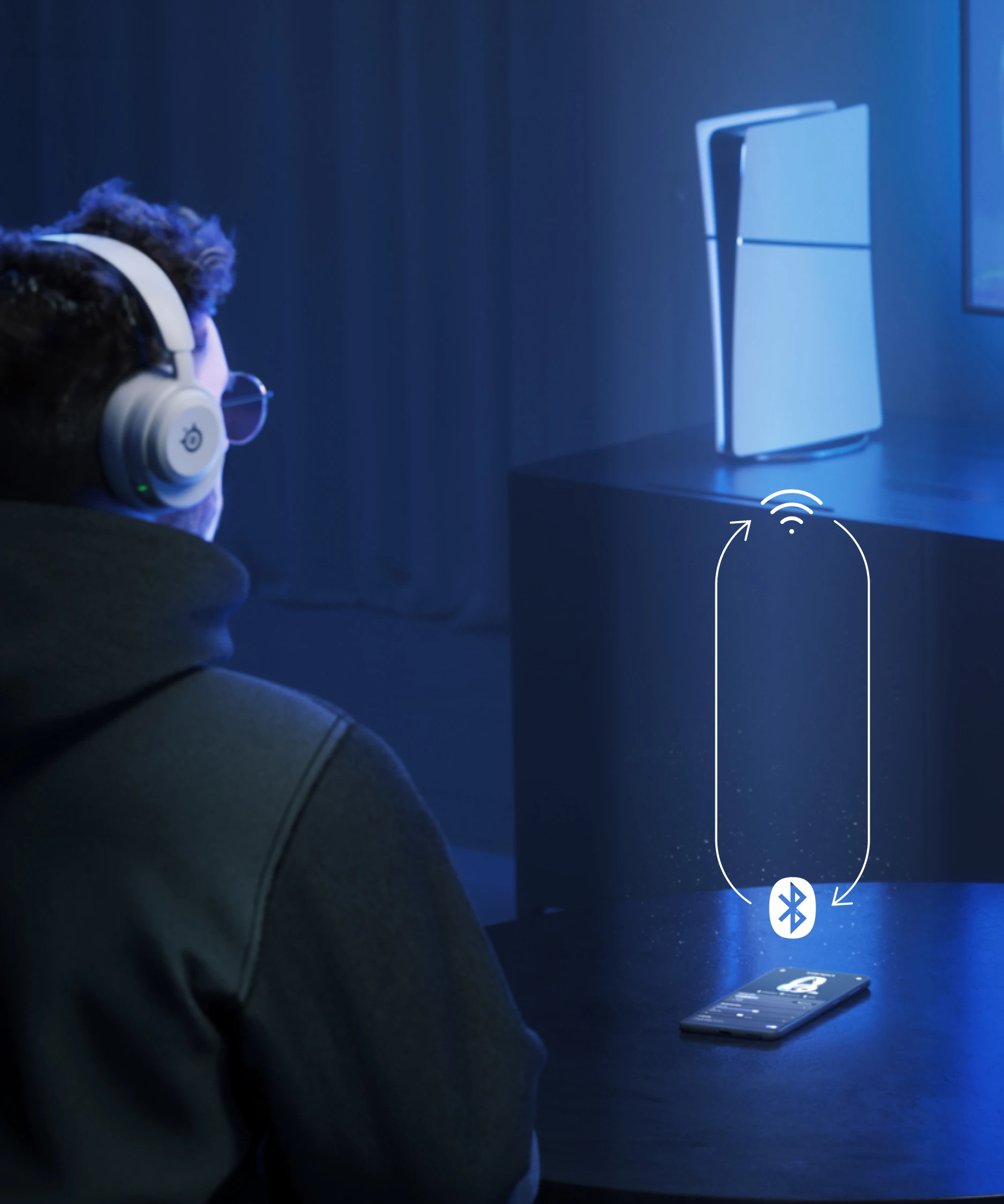 A man sitting on a couch with Arctis Nova 5 headphones on his ears; in front of him is a table with a phone on it. The man is looking at a distant TV, next to which stands a PS5 console.