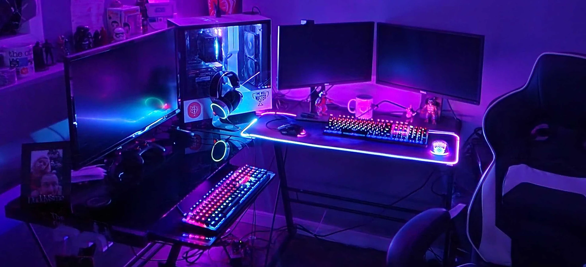 An expansive L-shaped gaming setup glowing with purple and pink RGB lighting, featuring triple monitors, two mechanical keyboards, a lit mousepad, and a white PC case with headphones.