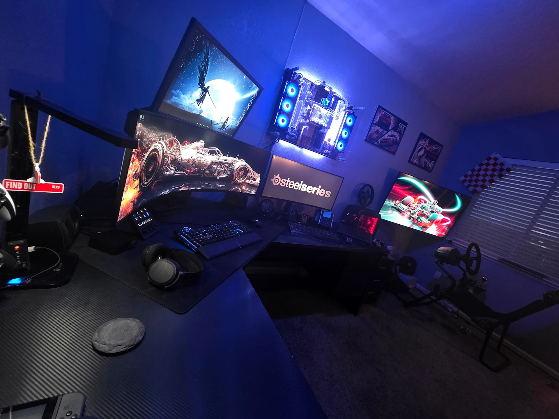 An expansive gaming setup glowing with blue RGB lighting, featuring four monitors and a curved screen, two mechanical keyboards, a lit mousepad, and a white PC case with headphones.