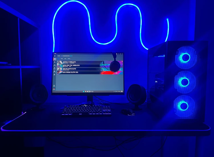 A high-tech gaming setup bathed in blue neon light, featuring a PC with glowing fans, a SteelSeries software menu on the monitor, and matching backlit peripherals on a dark desk.