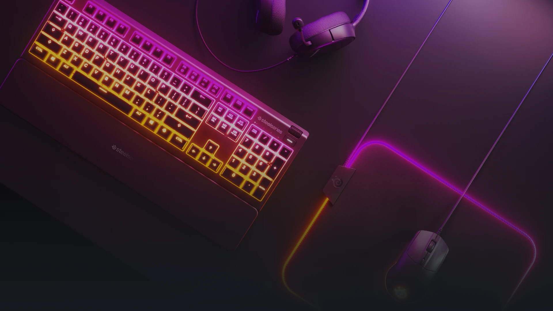  A top-down shot of a gaming keyboard, mouse, and headset on a dark surface. The keyboard and a large mousepad are illuminated with glowing yellow and purple lights.
