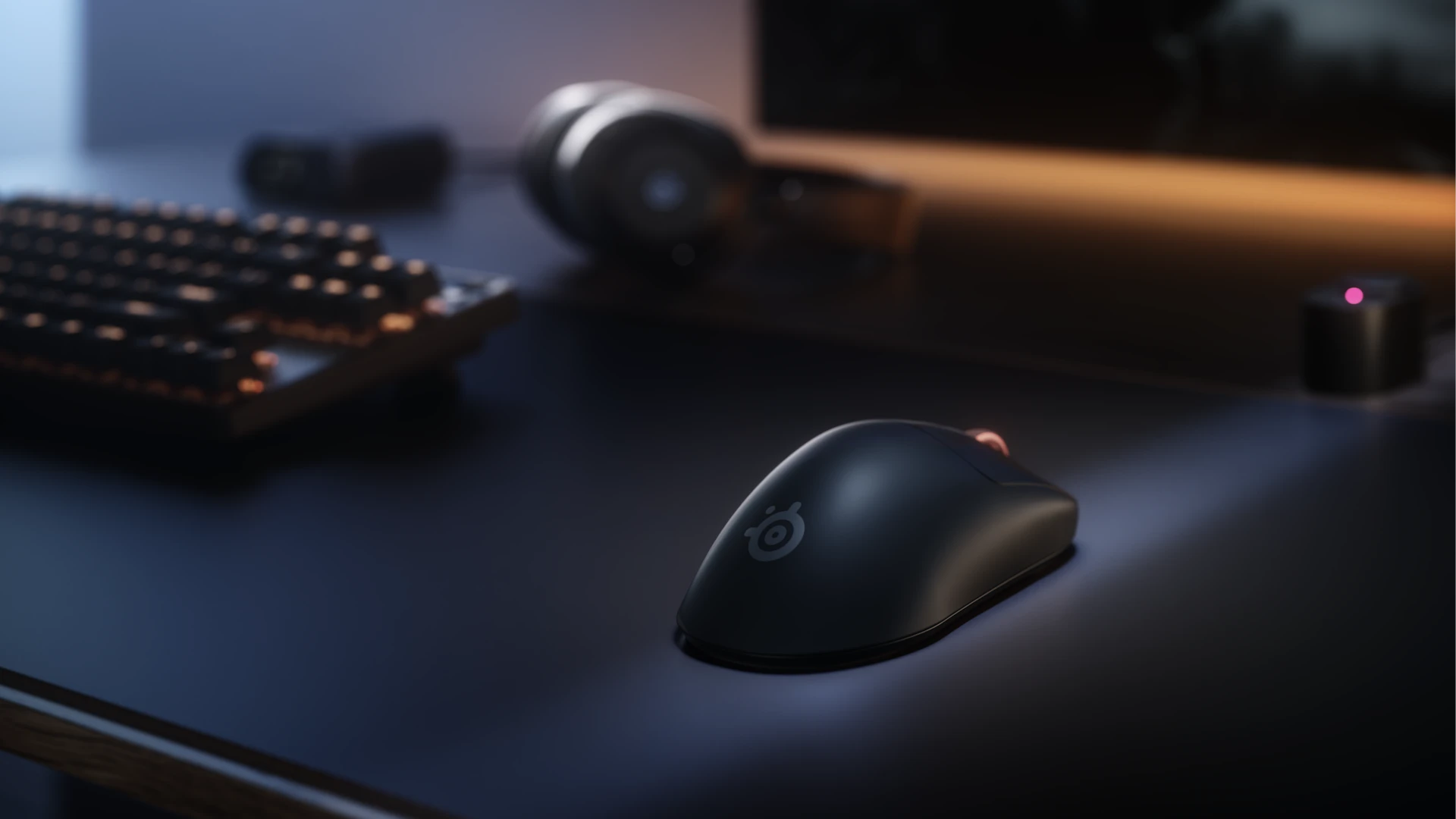 Šikmý pohled zezadu na matně černou herní myš Rival Pro ležící na stole vedle mechanické klávesnice a bezdrátových sluchátek.