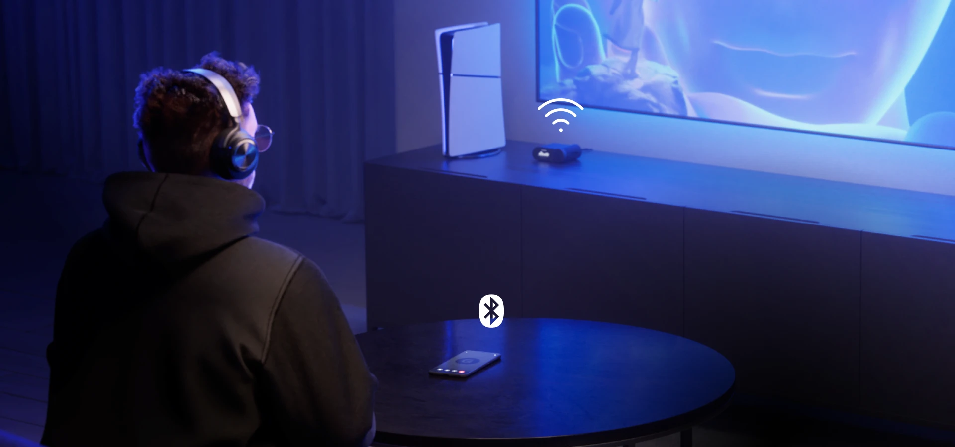 Un hombre sentado frente a un televisor grande con unos audífonos Arctis Nova Pro Wireless. Sobre la mesa frente a él hay un teléfono móvil, y en la mesita junto al televisor hay una consola y una estación de audífonos.