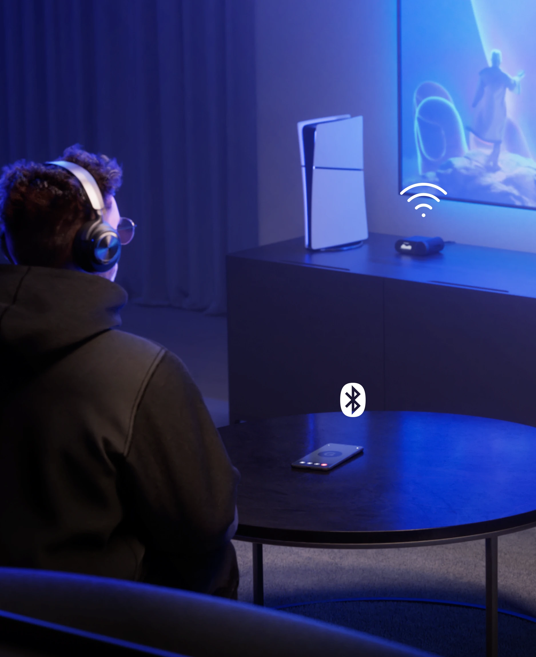 A man sitting in front of a large TV wearing Arctis Nova Pro Wireless headphones. On the table in front of him is a mobile phone, and on the side table next to the TV is a console and a headphone station
