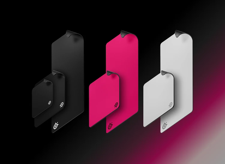 Uma linha de mousepads SteelSeries QcK em vários tamanhos (M a XXL) e cores: preto, rosa e branco. Cada mousepad apresenta uma dobra sutil no canto e o logotipo icônico.