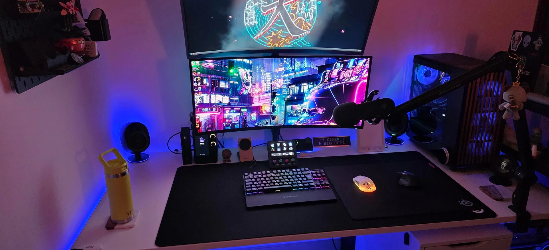 Stacked dual-monitor gaming setup with a cyberpunk wallpaper, a boom arm mic, Stream Deck, and mechanical keyboard on a large desk mat with vibrant blue ambient lighting.