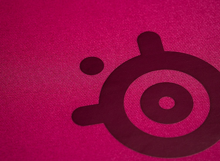 Primerísimo primer plano de la superficie de tela microtejida de alta densidad del SteelSeries QcK Heavy XXL en color magenta, optimizada para la precisión de seguimiento tanto de sensores ópticos como láser.