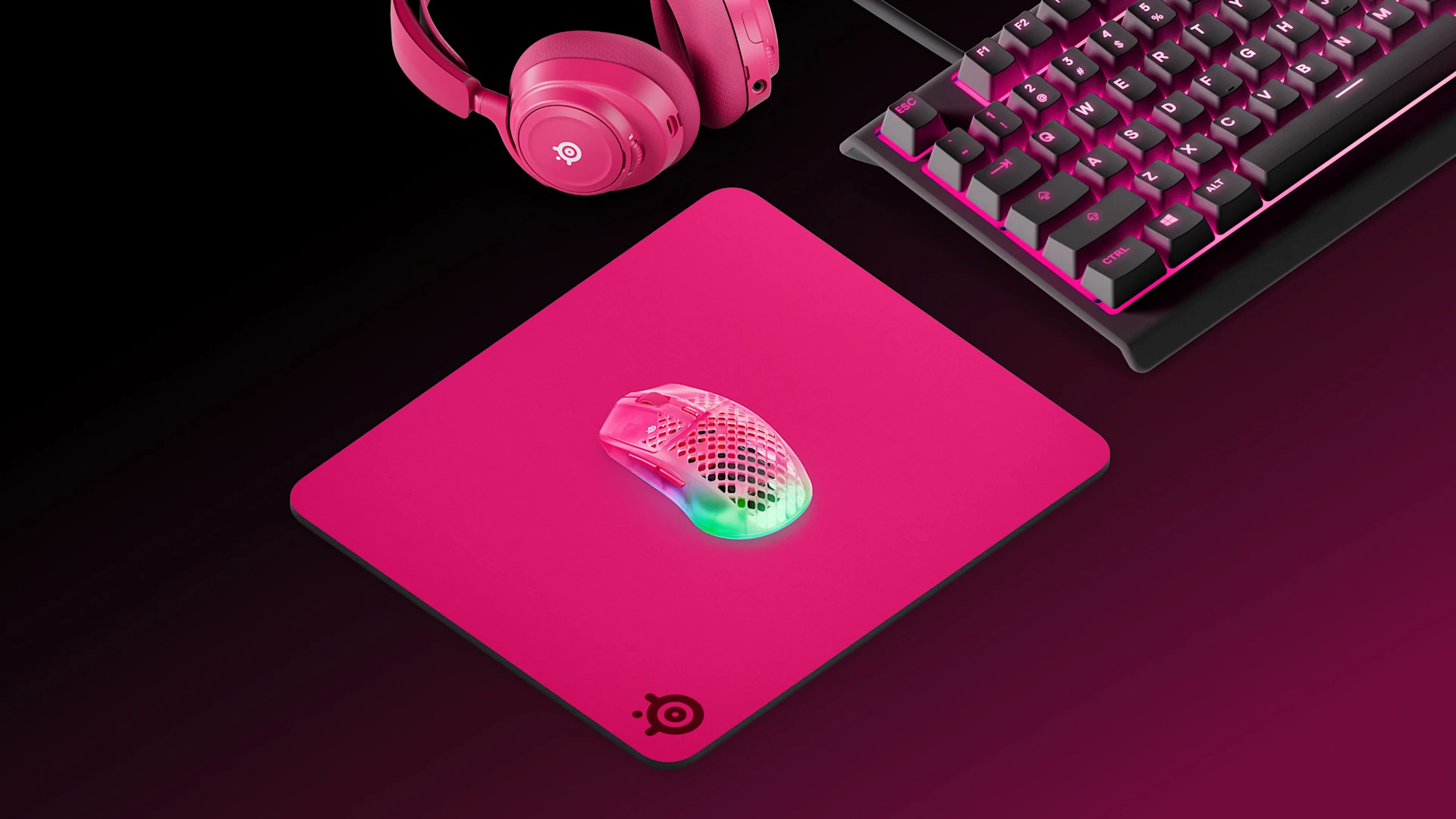 Isometrisk visning af et magenta SteelSeries-gaming-setup: QcK Heavy XXL-musemåtte, Apex 7 TKL-tastatur, Arctis Nova 7 magenta-headset og Aerox 3 Wireless-mus på en sort overflade.