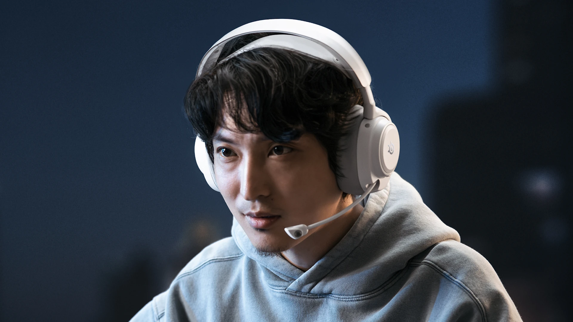Person wearing a light gray hoodie and white SteelSeries gaming headset with boom mic, focused indoors against a dark, softly blurred background.