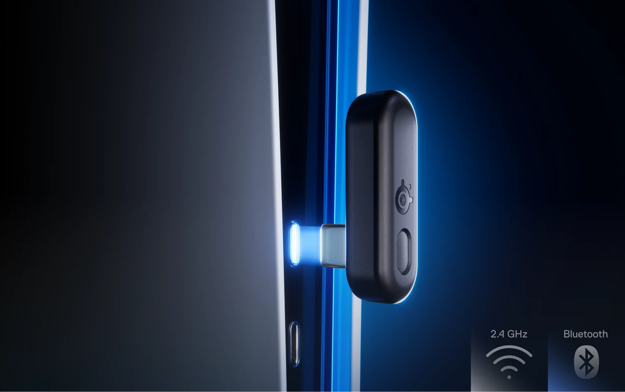 스틸시리즈 로고가 새겨진 작은 검은색 무선 USB-C 동글이 세련된 전자 기기에 꽂혀 파란색 불빛으로 빛나는 모습을 클로즈업한 모습입니다. 모서리에 있는 아이콘은 2.4GHz 무선 및 Bluetooth 연결을 지원함을 나타냅니다.