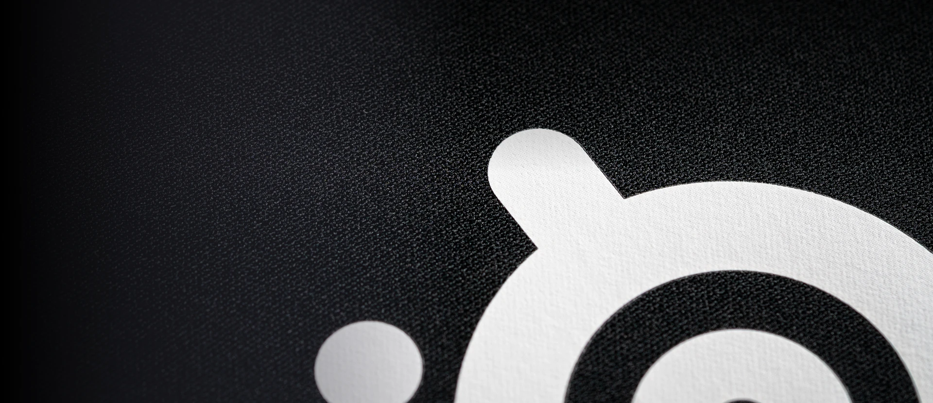 Primer plano en ángulo del logotipo blanco de SteelSeries sobre una superficie de tela microtejida negra, con el lado izquierdo difuminándose en una sombra profunda.