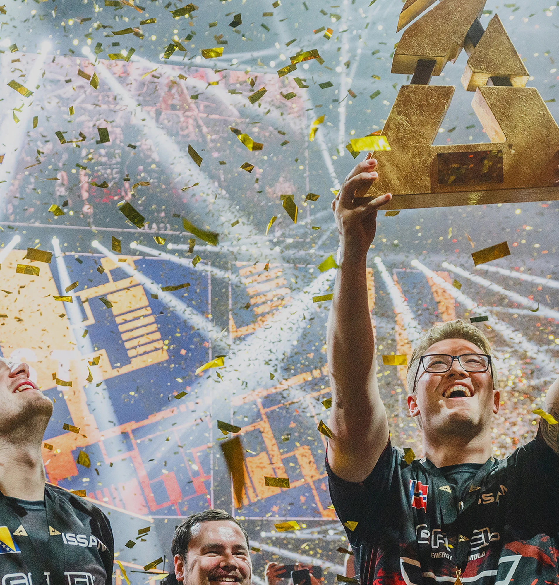 Two victorious esports players celebrate on a crowded stage under a shower of golden confetti, one joyfully hoisting a large, golden trophy.