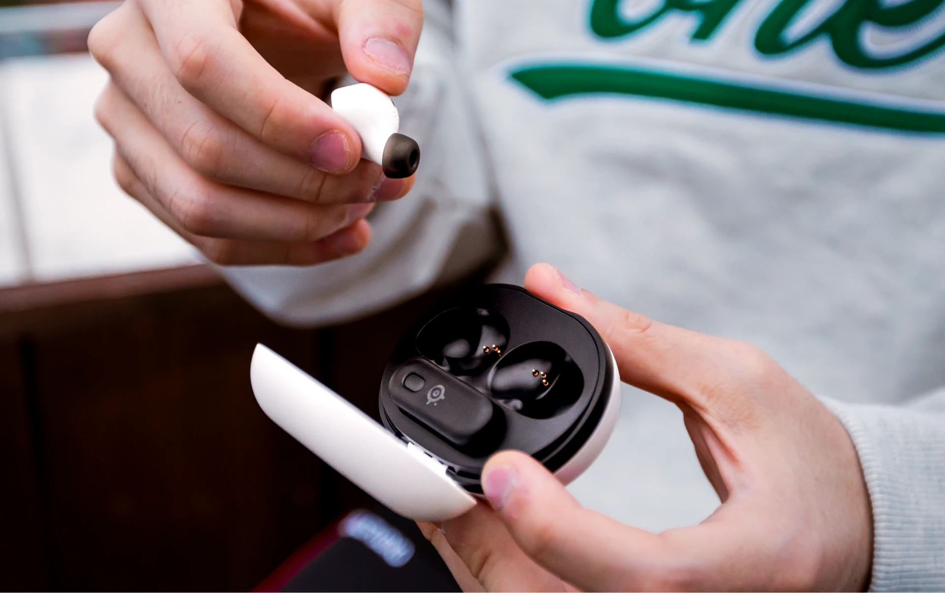Vista de los Arctis Gamebuds abiertos en una caja blanca; una persona los saca de la caja y sostiene un auricular en la mano.