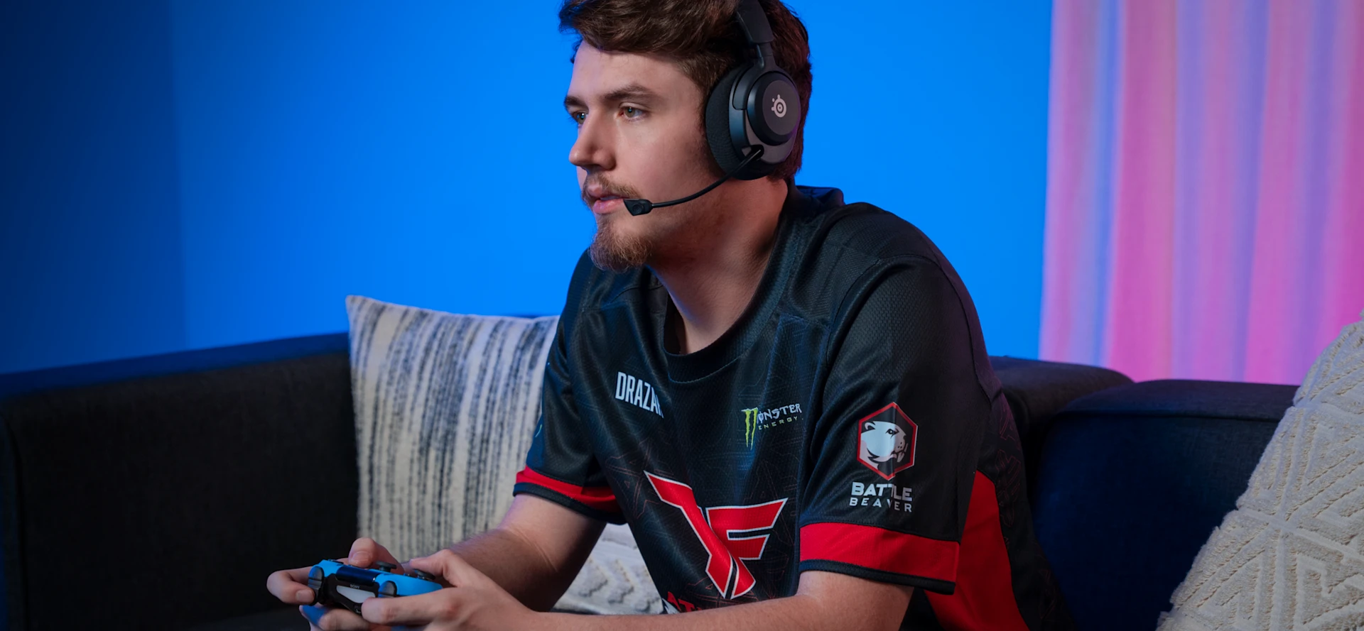 A young boy sitting on a couch wearing a FaZe t-shirt, holding a controller in his hands, and wearing Arctis Nova 5 headphones on his head.