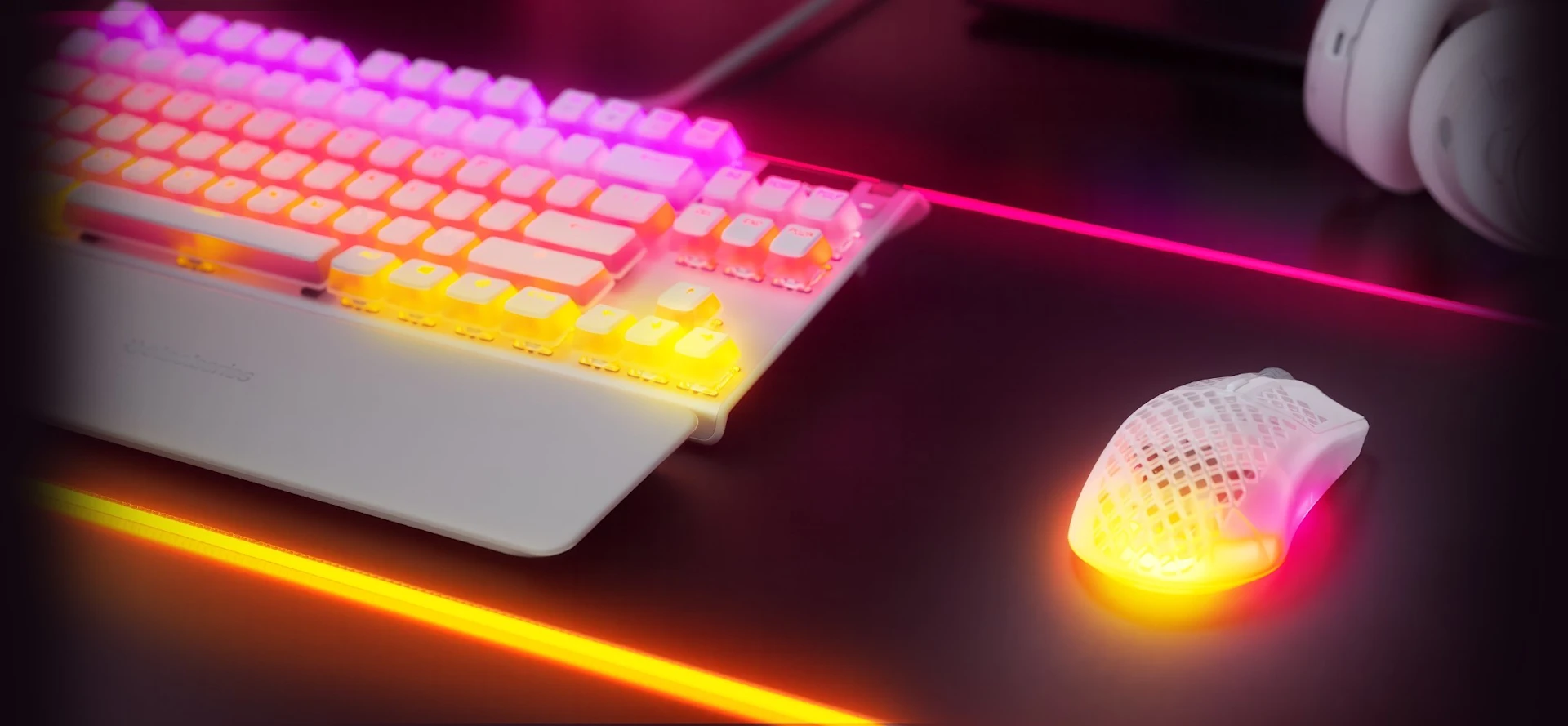  A close-up of a white gaming keyboard and a perforated mouse, both illuminated with vibrant pink and yellow RGB lighting on a dark desk.