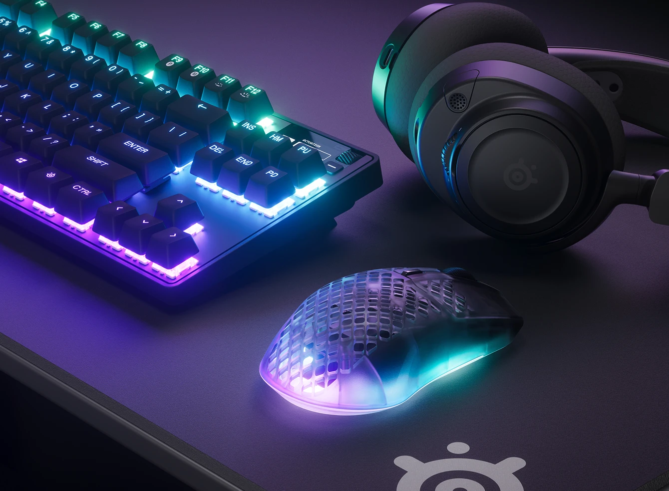 RGB keyboard, headset, and translucent gaming mouse on a desk in purple-blue lighting.