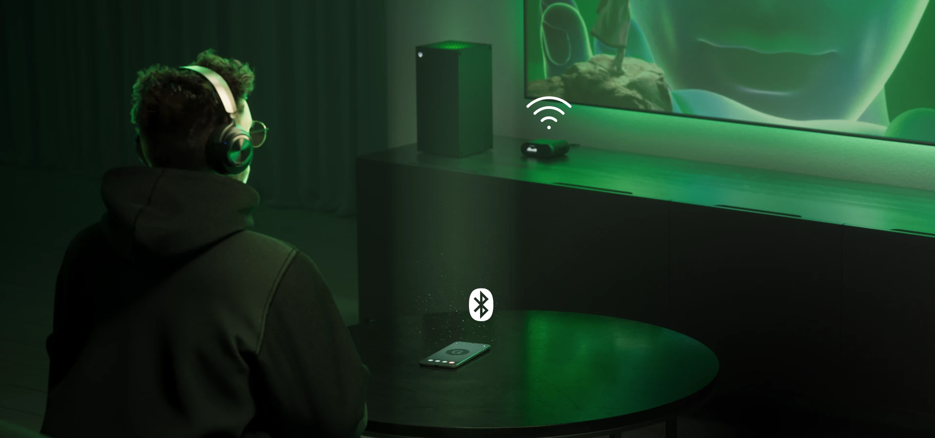 A man sitting in front of a large TV wearing Arctis Nova Pro Wireless headphones. On the table in front of him is a mobile phone, and on the side table next to the TV is a console and a headphone station