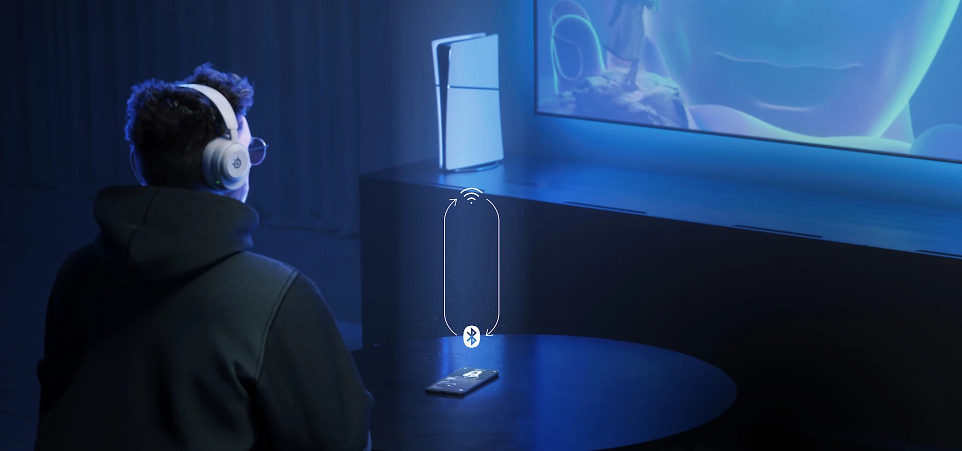 Un hombre sentado en un sofá con unos audífonos Arctis Nova 5 en los oídos; frente a él hay una mesa con un teléfono. El hombre mira un televisor lejano, junto al cual se encuentra una consola PS5.