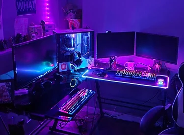 An expansive L-shaped gaming setup glowing with purple and pink RGB lighting, featuring triple monitors, two mechanical keyboards, a lit mousepad, and a white PC case with headphones.