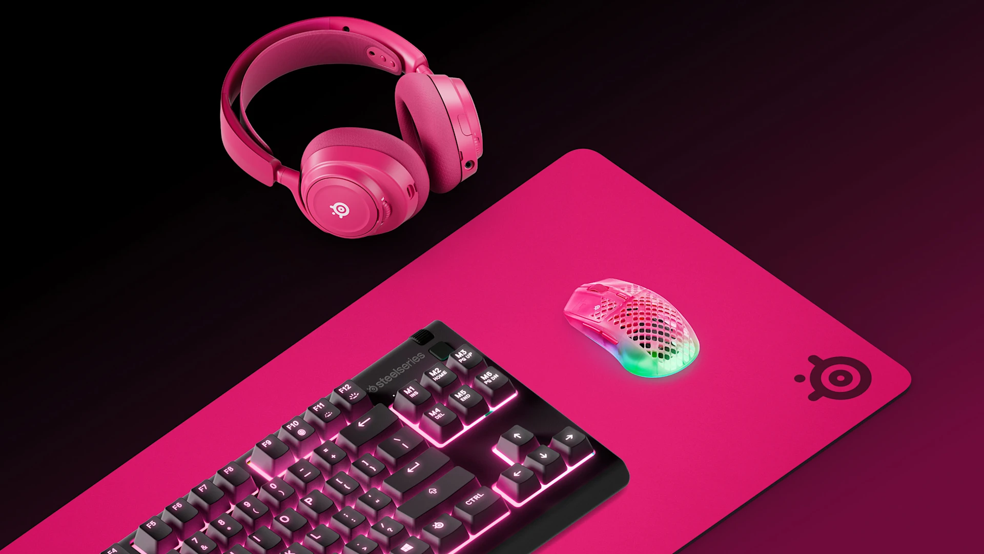 Vista isométrica de una configuración de SteelSeries en color magenta: alfombrilla QcK Heavy XXL, teclado Apex 7 TKL, auriculares Arctis Nova 7 y ratón Aerox 3 sobre una superficie negra.
