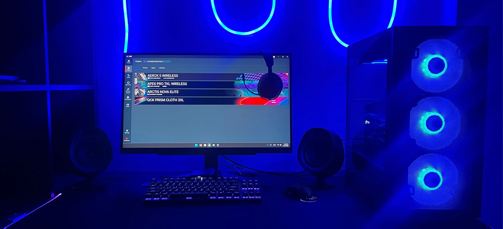 A high-tech gaming setup bathed in blue neon light, featuring a PC with glowing fans, a SteelSeries software menu on the monitor, and matching backlit peripherals on a dark desk.