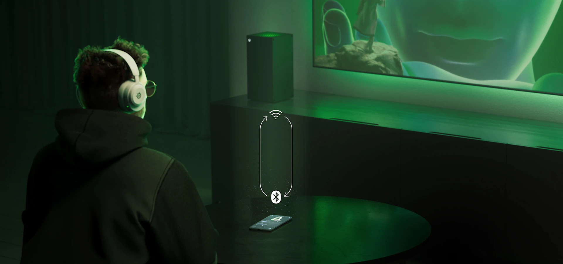 Un hombre sentado en un sofá con unos audífonos Arctis Nova 5 en los oídos; frente a él hay una mesa con un teléfono. El hombre mira un televisor lejano, junto al cual se encuentra una consola PS5.