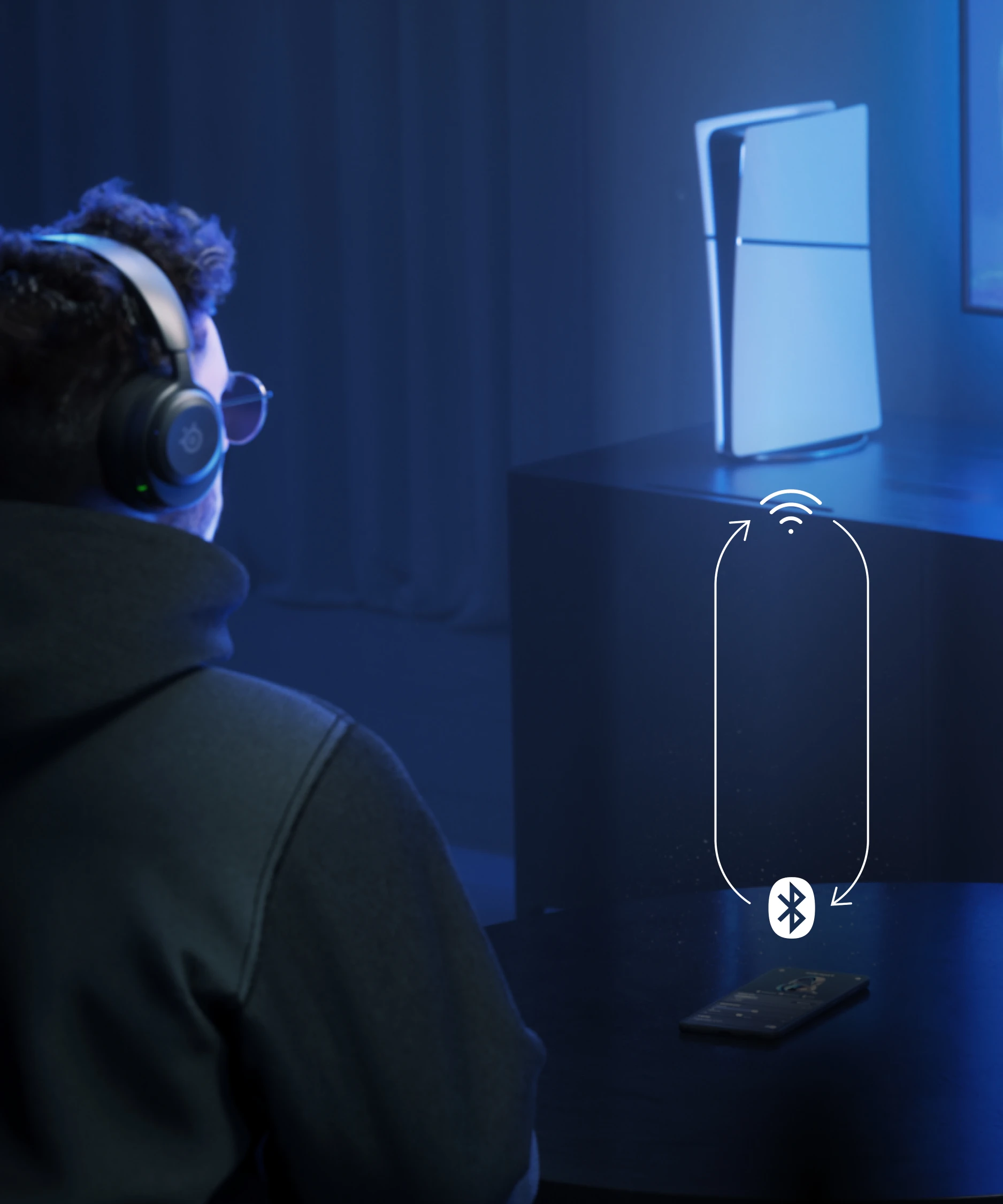 A man sitting on a couch with Arctis Nova 5 headphones on his ears; in front of him is a table with a phone on it. The man is looking at a distant TV, next to which stands a PS5 console.