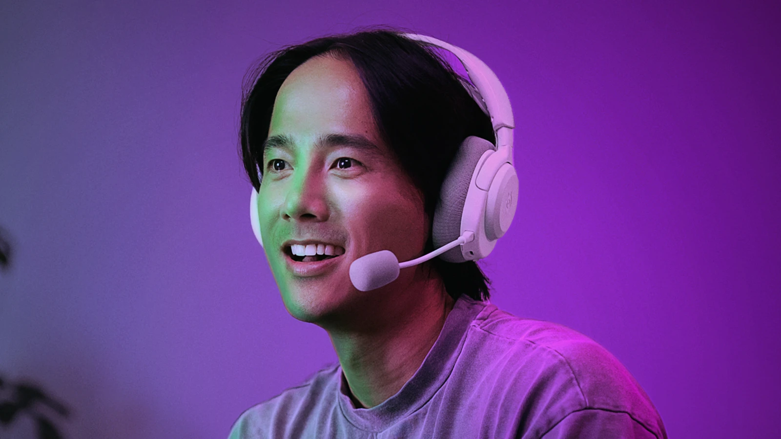 A gamer wearing the white SteelSeries Arctis Nova 3 Wireless headset with the microphone extended, smiling against a vibrant purple and green atmospheric background.