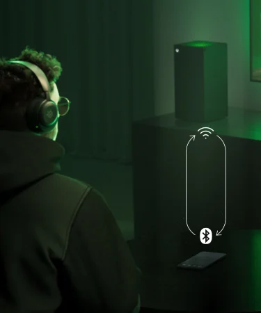 A man sitting on a couch with Arctis Nova 5 headphones on his ears; in front of him is a table with a phone on it. The man is looking at a distant TV, next to which stands a PS5 console.