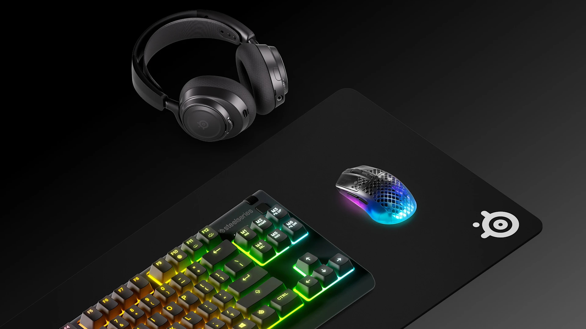 High-angle view of a SteelSeries gaming setup: RGB mechanical keyboard, wireless mouse, and headset on a large black QcK mousepad. Balanced professional layout on a dark background.