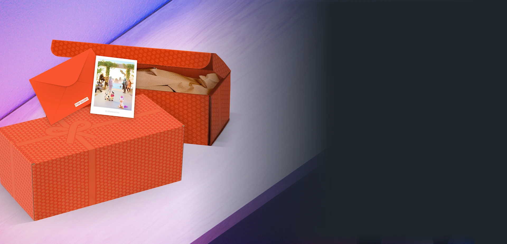 Two orange SteelSeries gift boxes, one closed with a printed bow and one open with brown packing paper, next to an orange envelope and a small polaroid photo on a light surface.