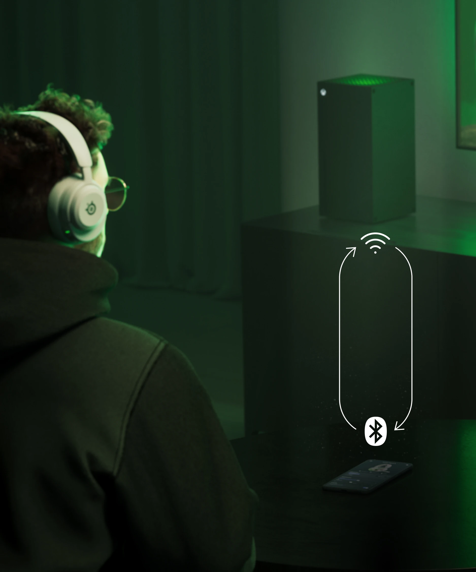 A man sitting on a couch with Arctis Nova 5 headphones on his ears; in front of him is a table with a phone on it. The man is looking at a distant TV, next to which stands a PS5 console.