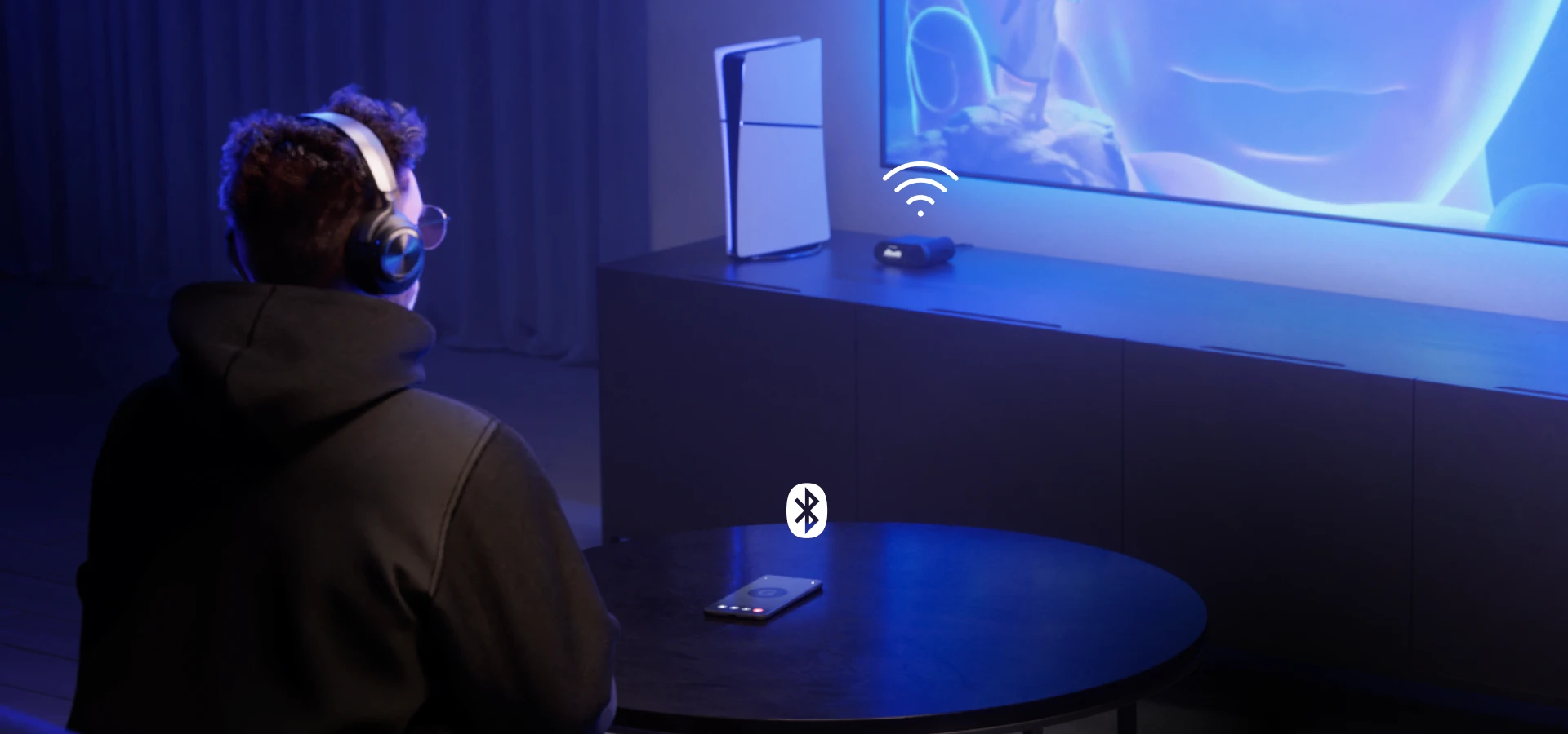 A man sitting in front of a large TV wearing Arctis Nova Pro Wireless headphones. On the table in front of him is a mobile phone, and on the side table next to the TV is a console and a headphone station