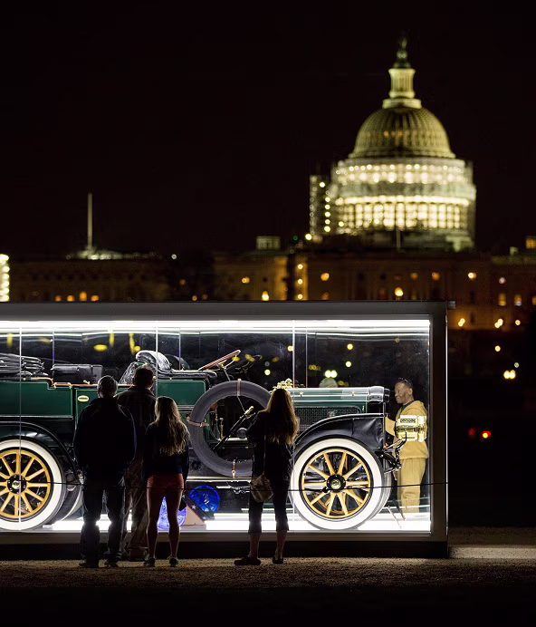 People view a vintage car displayed in a glass case at night with the U.S. Capitol building illuminated in the background.